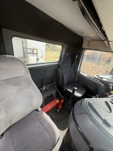Cab interior of a 2005 Volvo VNL64T300 truck showing driver seat, passenger seat, steering wheel, dashboard, gear lever, and overhead compartments.