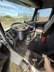 Cab interior of a Volvo truck showing steering wheel, dashboard, pedals, and driver's seat with a hand on a grip; window with writing on glass and desert landscape outside.