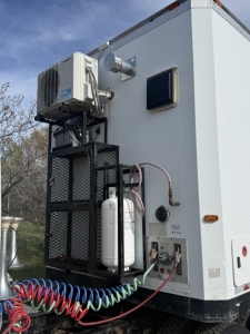 Truck utility area with external A/C condenser, white refrigerator unit, hoses, and control panel on a Volvo VNL trailer unit; visible pipes, wires, and a small window.