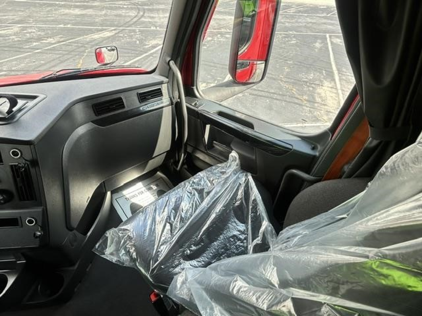 Cab interior of a Freightliner Cascadia truck showing driver's seat and dashboard with plastic-covered passenger seat and window view.