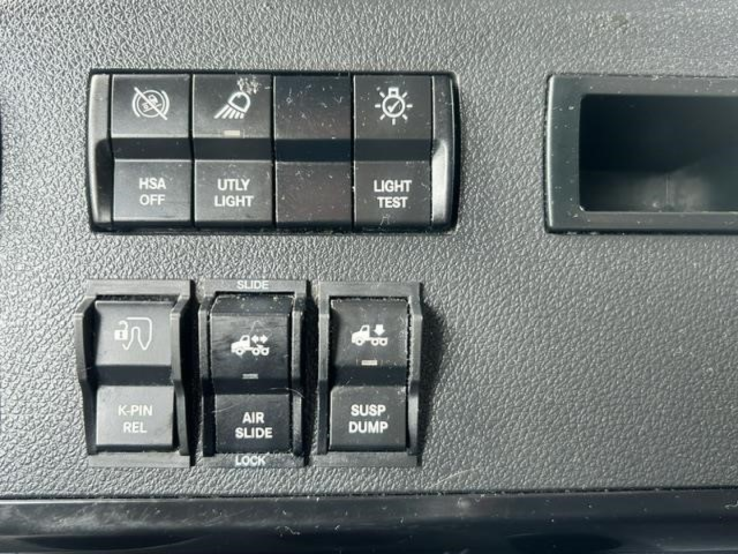 Dashboard controls of a Freightliner Cascadia showing switches labeled HSA OFF, UTLY LIGHT, LIGHT TEST, K-PIN REL, AIR SLIDE, SUSP DUMP, with a storage compartment on the right.