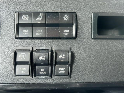 Dashboard controls of a Freightliner Cascadia showing switches labeled HSA OFF, UTLY LIGHT, LIGHT TEST, K-PIN REL, AIR SLIDE, SUSP DUMP, with a storage compartment on the right.