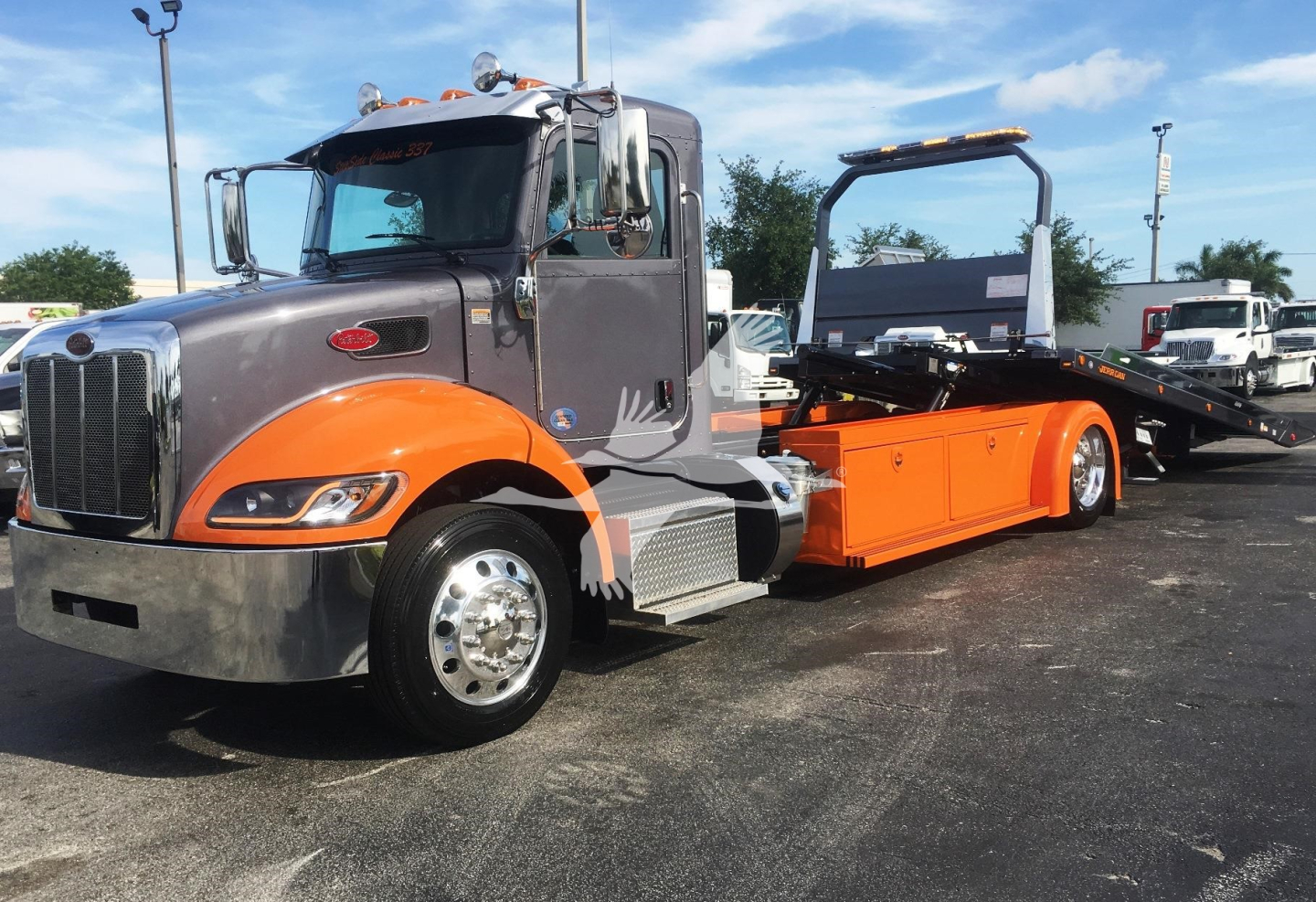 Peterbilt 337 rollback wrecker truck in gray and orange, front cab view, low angle, with flatbed and winch apparatus in background.