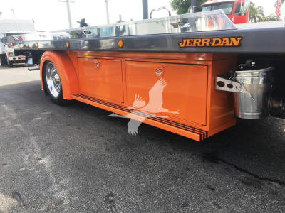 Orange Jerr-Dan rollback service body with storage compartments and gray diamond-plate bed edge. JERR-DAN logo on side; metal canister near rear. Wheel and trailer hitch area visible.
