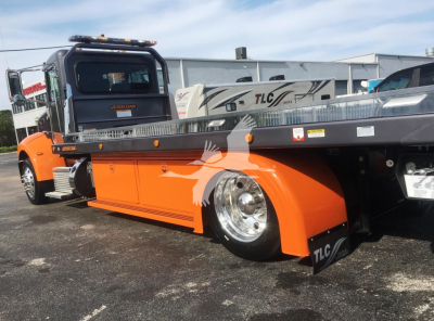 Peterbilt 337 rollback truck showing orange fenders and flatbed; visible text includes TLC signage on fender, and various decals along the truck bed and cab window.