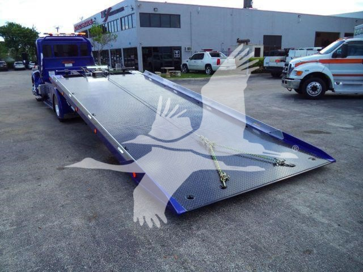 Blue Peterbilt rollback truck at a dealership with a long flatbed tow bed extended on a white and blue tow truck; other vehicles and a building in the background.