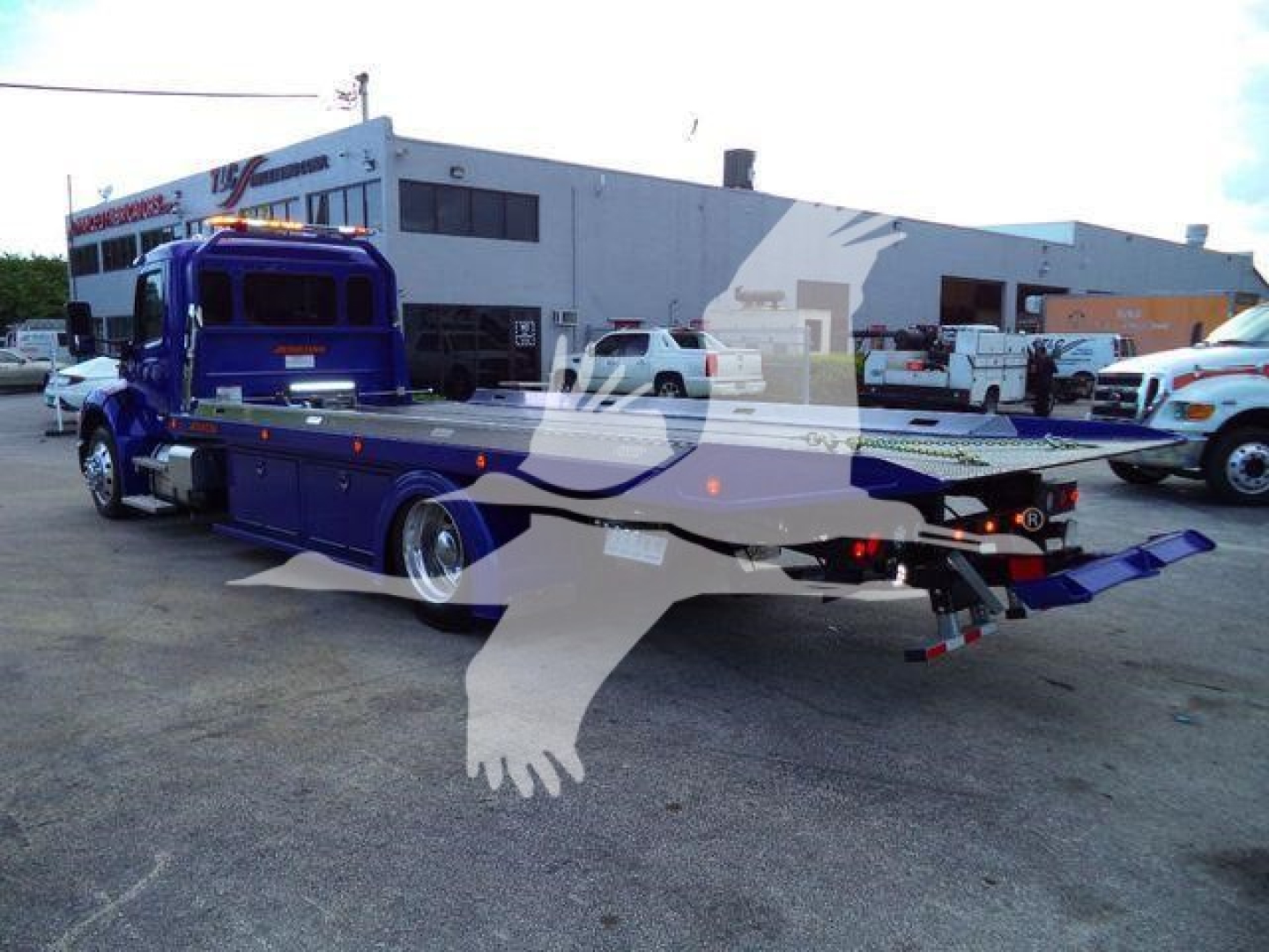BluePeterbilt rollback truck, flatbed, side view with storage compartments and rear ramp extended; dealership lot in background.