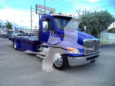 Blue 2025 Peterbilt 536 rollback truck in a parking lot, front grille, chrome bumper, and flatbed with side rails visible.