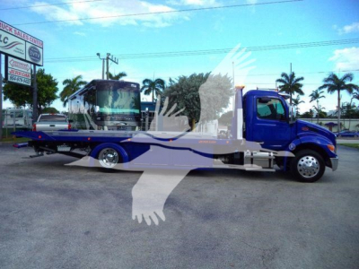 2025 Peterbilt 536 rollback truck in blue and gray, flatbed with hydraulic equipment visible, side view with branding and palm trees in background.