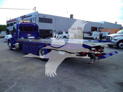 BluePeterbilt rollback truck, flatbed, side view with storage compartments and rear ramp extended; dealership lot in background.