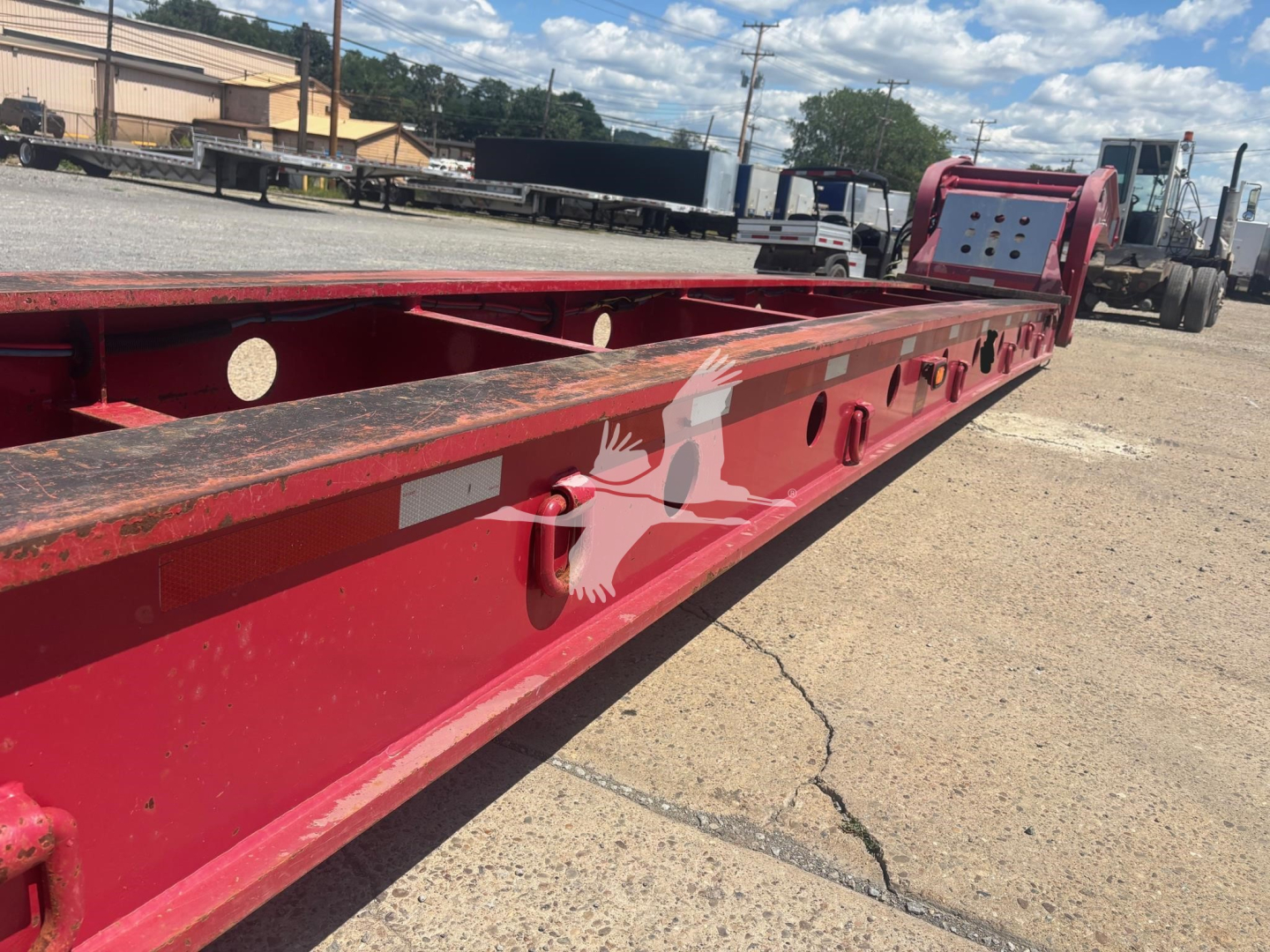 Lowboy trailer frame showing red structural rails with welded braces and circular cutouts along the side, background includes trucks and lot.