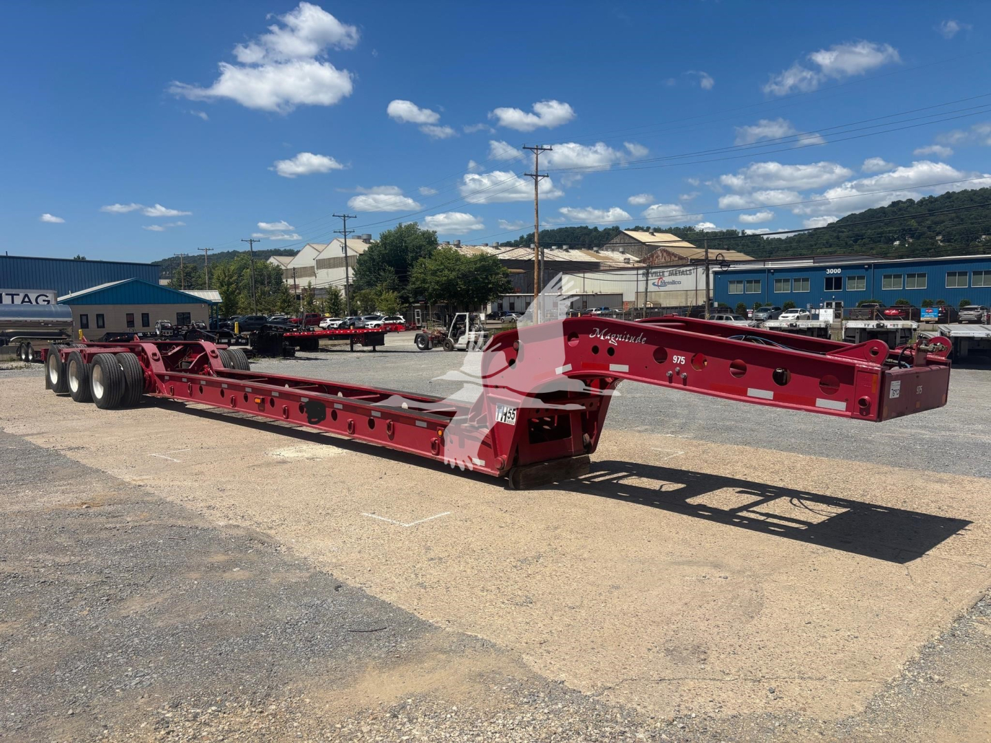Flatbed beam trailer with red-painted structure and multiple axles in an outdoor lot; visible hydraulic and support components along the frame.