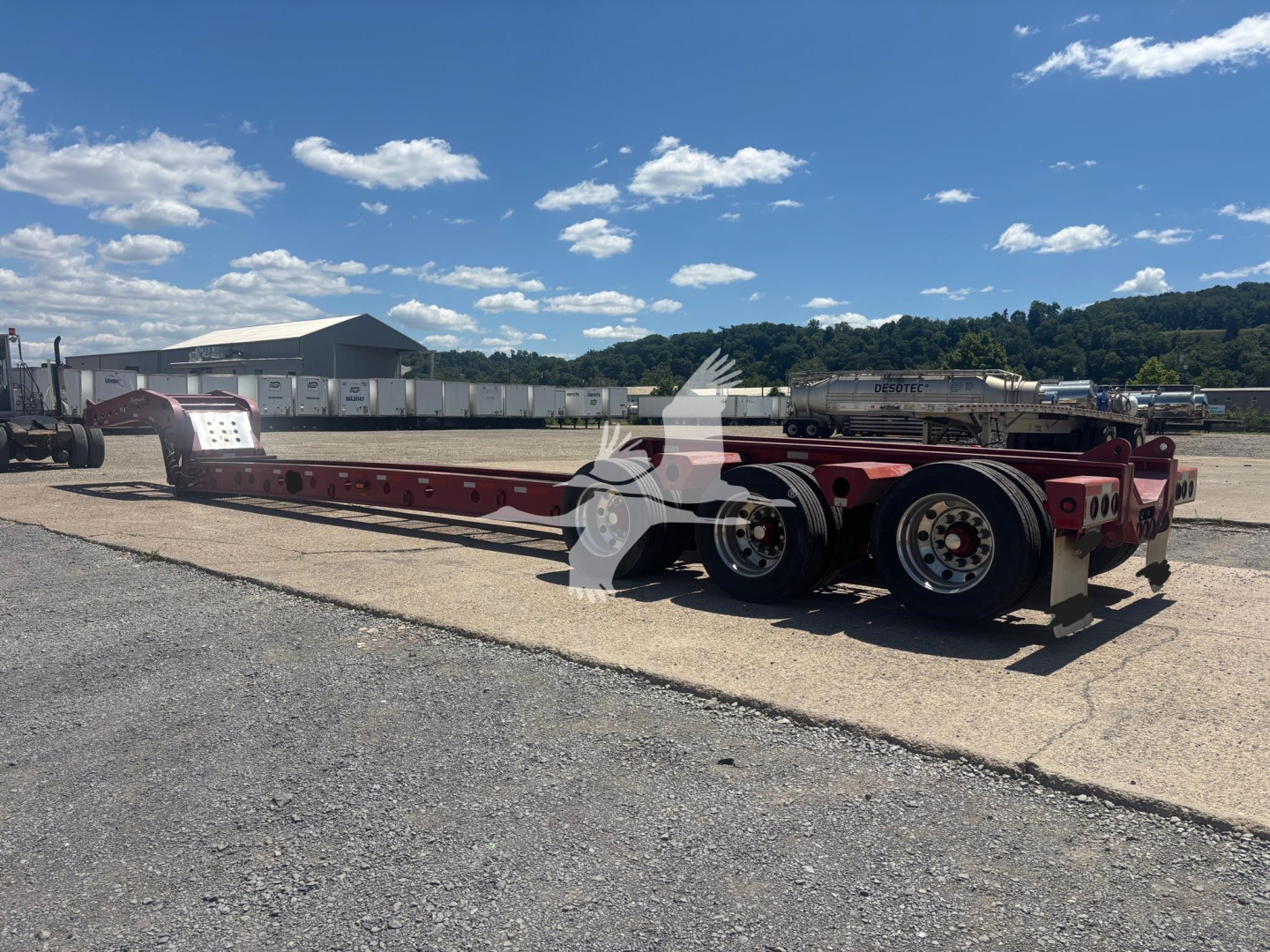Lowboy trailer with red frame and dual rear axles, flatbed beam showing empty deck, with clear sky and industrial yard in background.