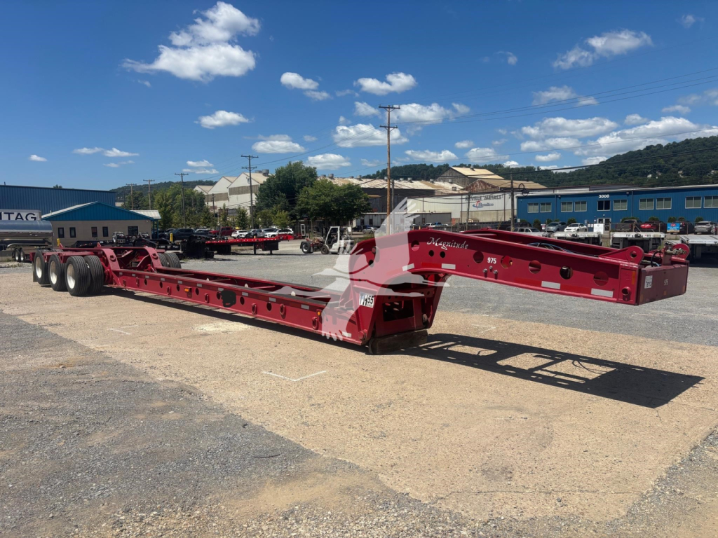 Flatbed beam trailer with red-painted structure and multiple axles in an outdoor lot; visible hydraulic and support components along the frame.