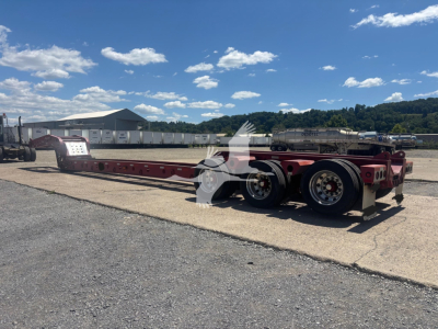 Lowboy trailer with red frame and dual rear axles, flatbed beam showing empty deck, with clear sky and industrial yard in background.