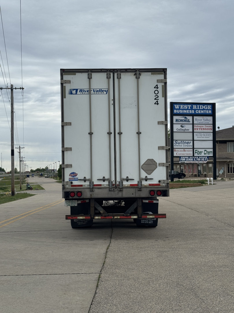 Rear view of a white utility trailer with doors; visible numbers 4024 on the right door, small diamond placard, and a blue River Valley logo sticker on the left door. Street and business signs in distance.