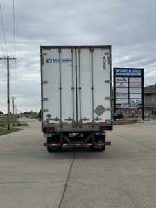 Rear view of a white utility trailer with doors; visible numbers 4024 on the right door, small diamond placard, and a blue River Valley logo sticker on the left door. Street and business signs in distance.