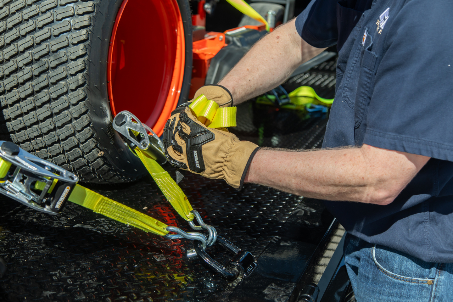 Isuzu NQR flatbed truck being secured with yellow tie-down straps to a trailer; close-up of a mechanic’s gloved hands attaching strap to a wheel and hook.