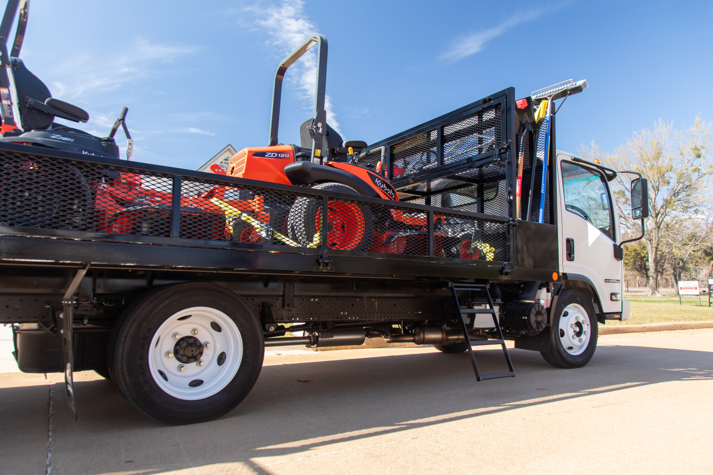 Isuzu NQR flatbed tow truck with open bed carrying a small orange ride-on mower; side railing, metal ladder, and cab visible.
