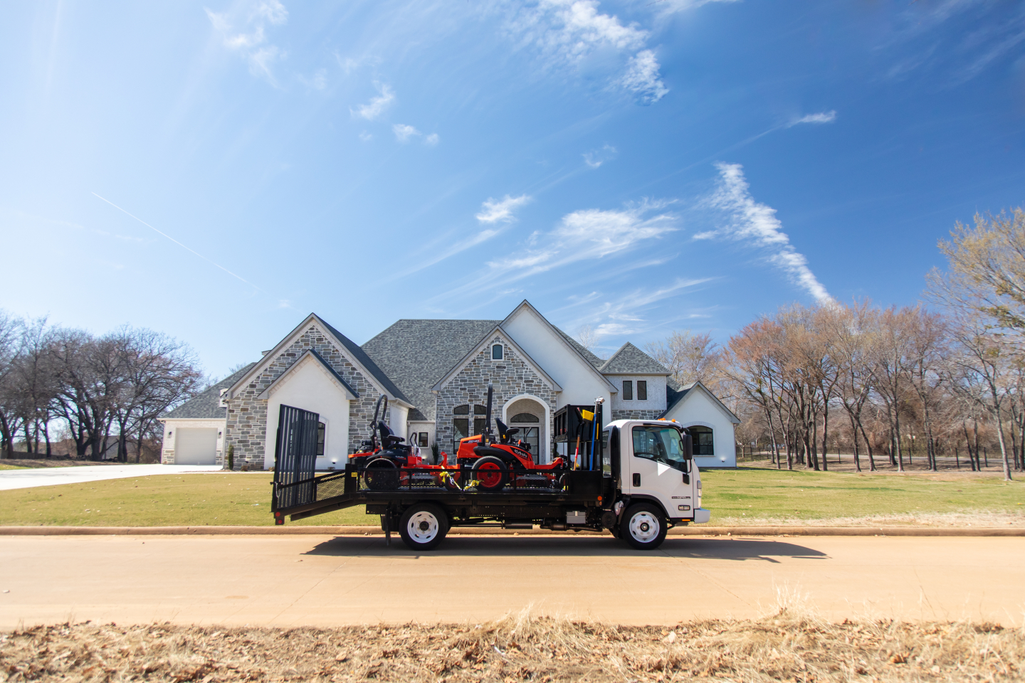 Isuzu flatbed truck with a small red tractor loaded on a tiltbed, parked in front of a large house with a stone facade under a clear blue sky.