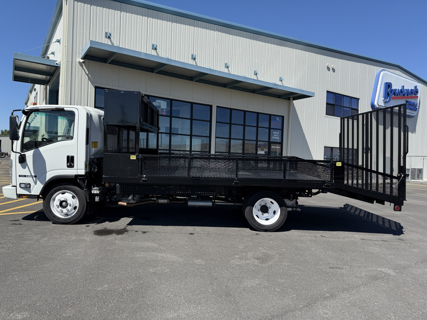 Isuzu NQR flatbed truck with a loading ramp and utility bed in a dealership lot, side view showing cab and cargo area.