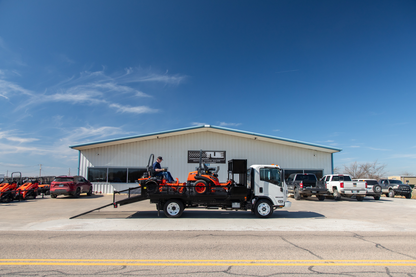 Isuzu NQR flatbed truck in a lot with a ramp and a small crane on the bed, vehicle and equipment visible in front of a light warehouse building.
