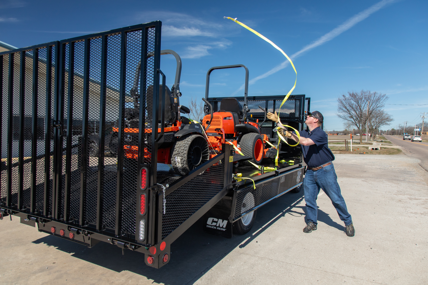 Isuzu flatbed truck loaded on trailer with utility equipment and a man playing with a long yellow strap in an outdoor lot.