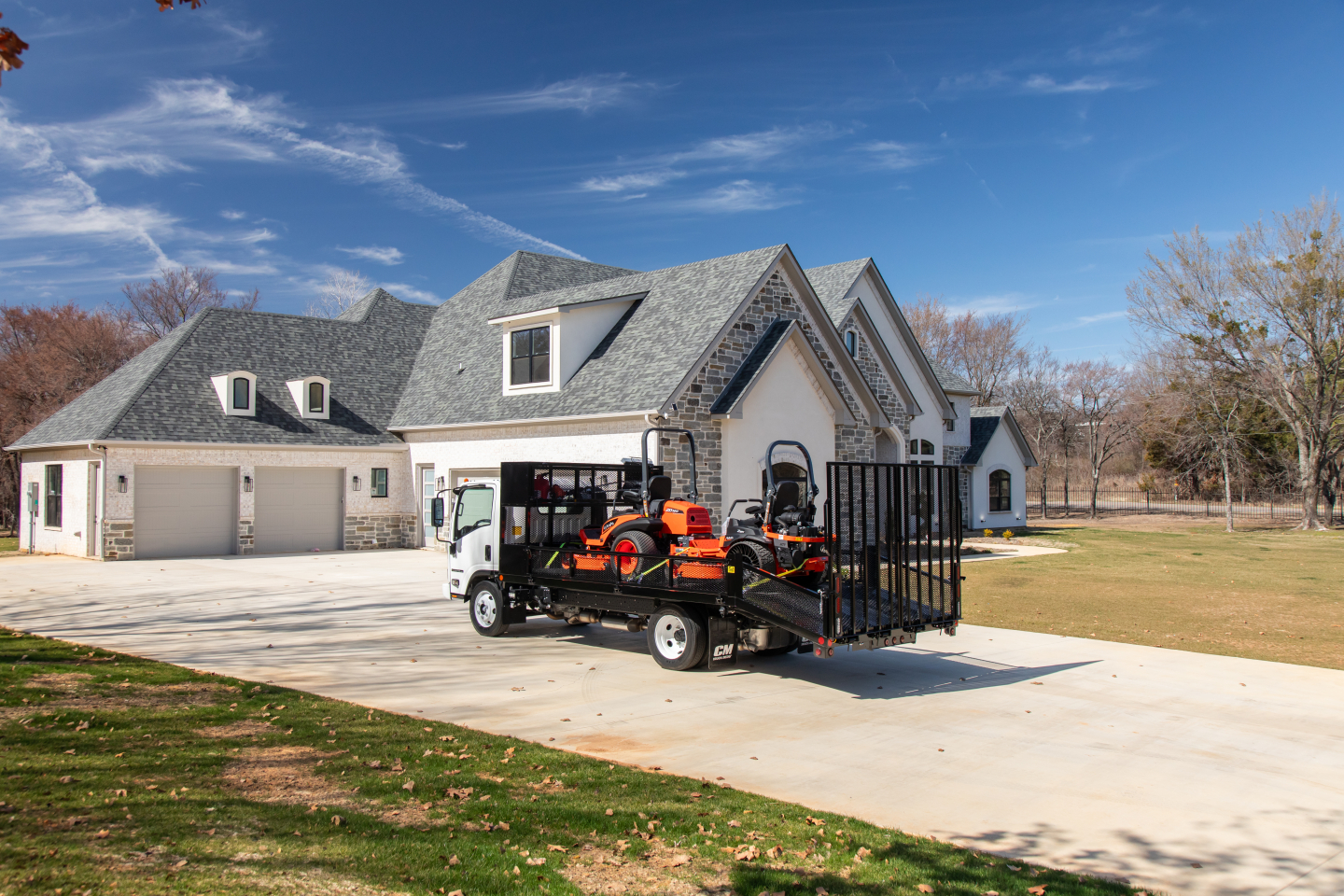 Isuzu flatbed truck with equipment on trailer in front of a large house, clear blue sky, residential driveway.