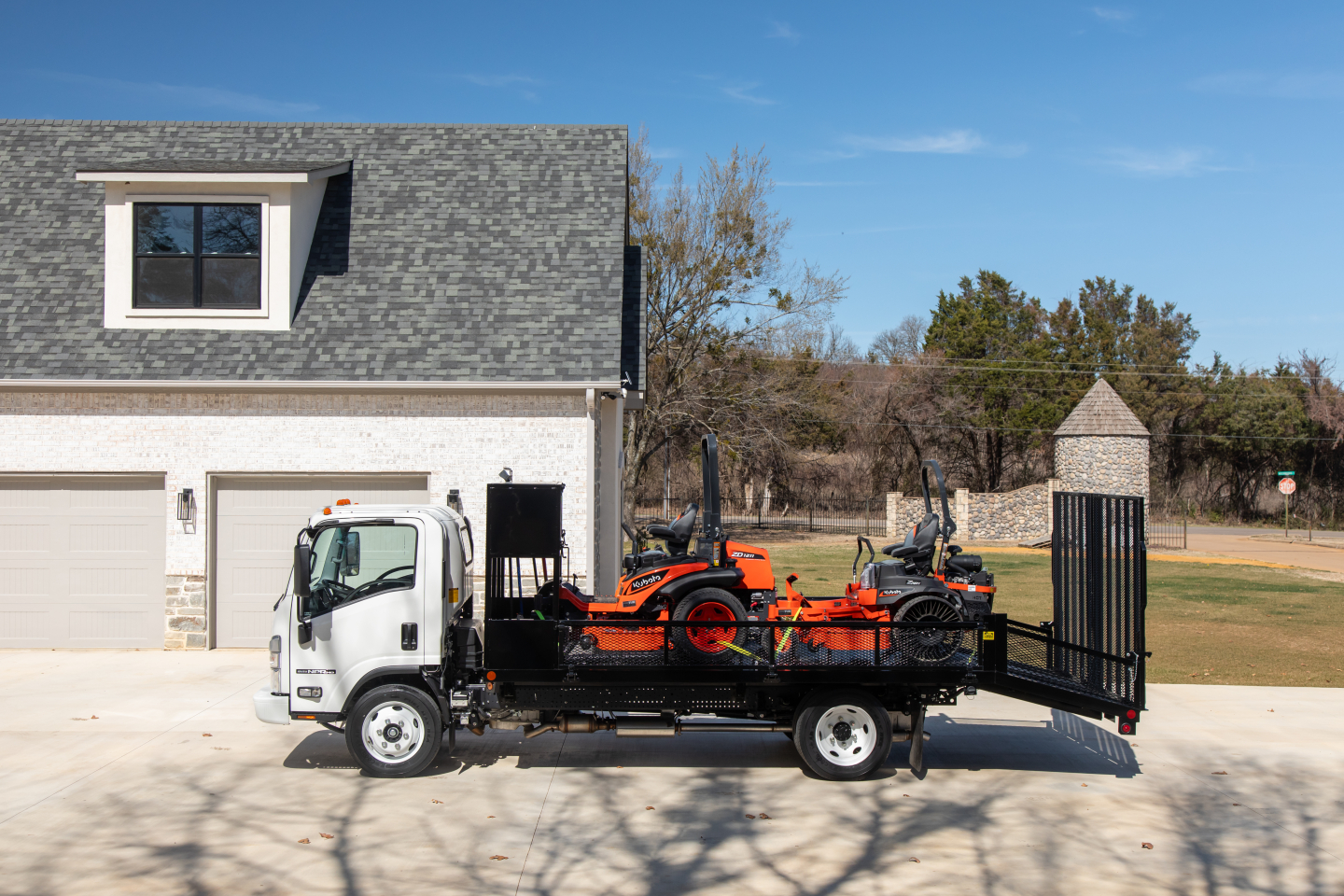 Isuzu flatbed truck with a car carrier loading two orange and black riding lawn mowers on a trailer, white cab, residential driveway, clear sky.