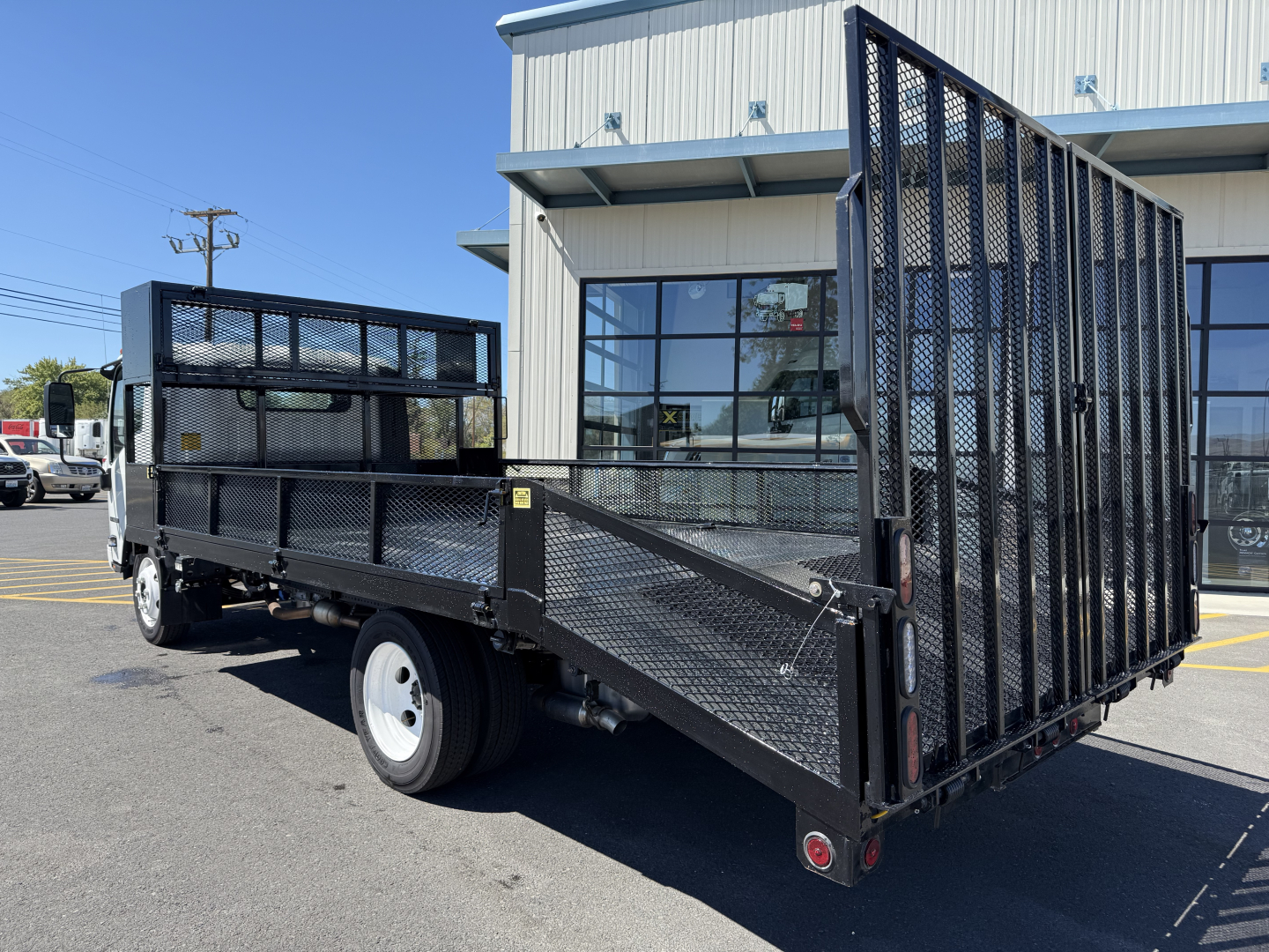 Isuzu flatbed truck trailer in a dealership lot, black metal mesh dump gate in rear, cargo area open, left side view