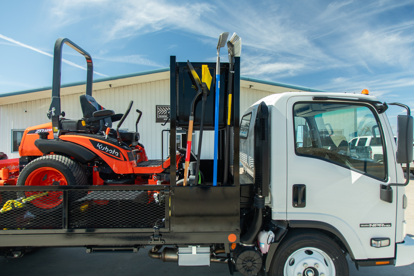 Isuzu NQR-GAS flatbed truck with a Kubota tractor on the flatbed carrying shovels and equipment visible in the truck bed.