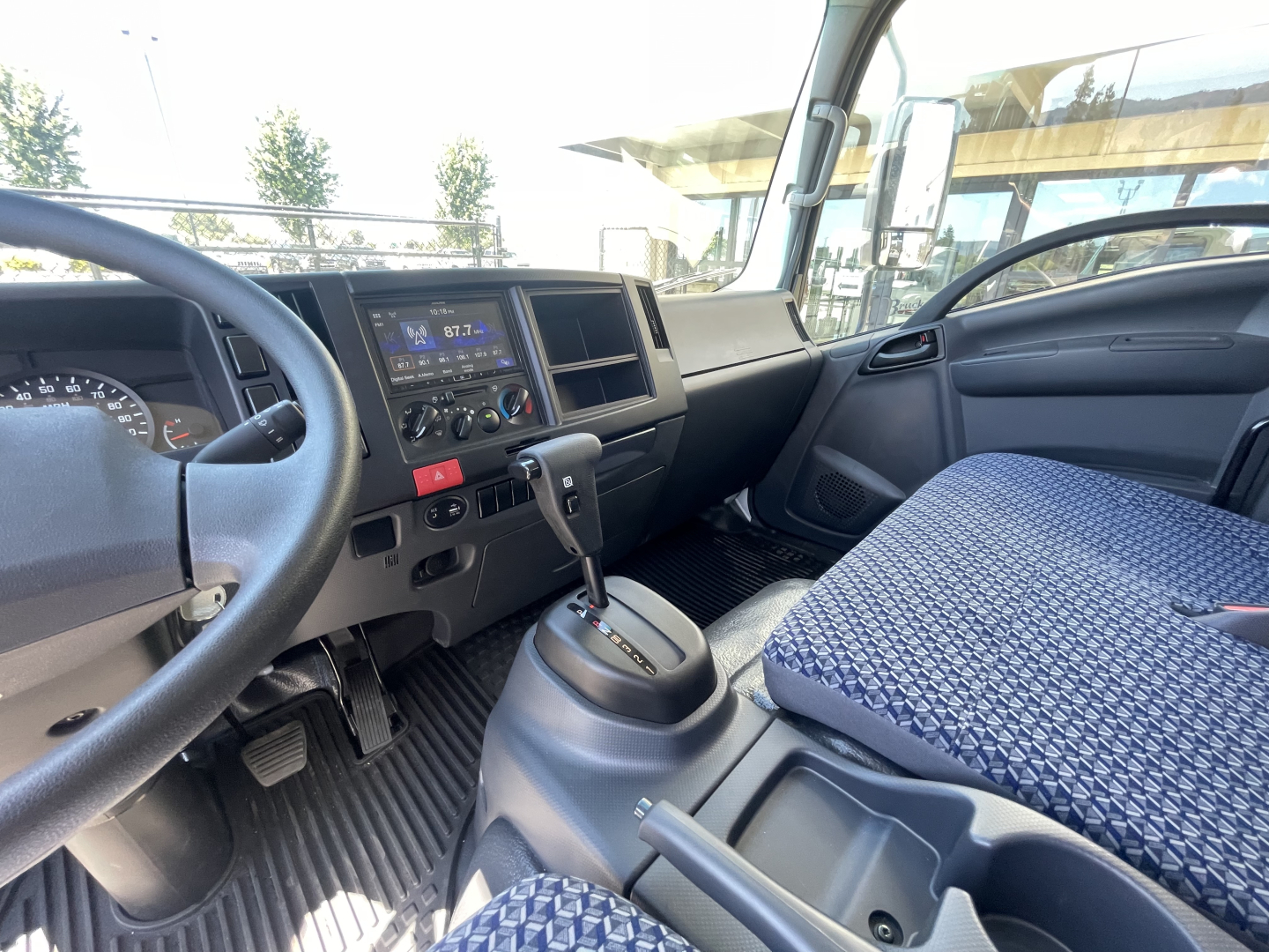 Isuzu NQR cab interior showing steering wheel, dashboard with digital radio display, climate controls, gear shifter, and blue patterned driver seat.