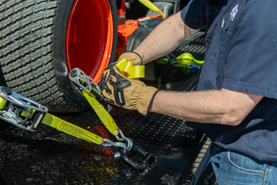 Isuzu NQR flatbed truck being secured with yellow tie-down straps to a trailer; close-up of a mechanic’s gloved hands attaching strap to a wheel and hook.