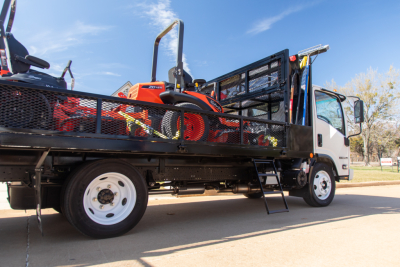 Isuzu NQR flatbed tow truck with open bed carrying a small orange ride-on mower; side railing, metal ladder, and cab visible.