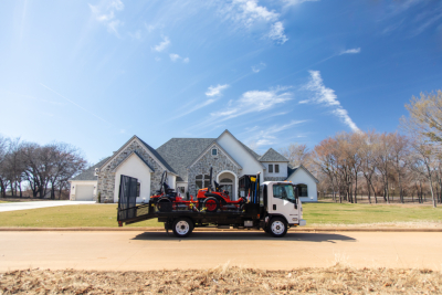 Isuzu flatbed truck with a small red tractor loaded on a tiltbed, parked in front of a large house with a stone facade under a clear blue sky.