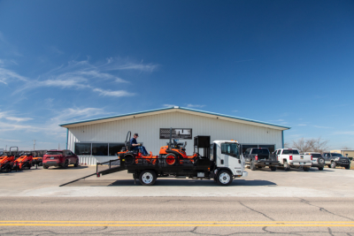 Isuzu NQR flatbed truck in a lot with a ramp and a small crane on the bed, vehicle and equipment visible in front of a light warehouse building.