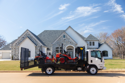 Isuzu flatbed truck with a load of small tractors on a black flatbed, white cab, residential street and blue sky in background.