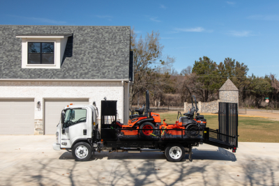 Isuzu flatbed truck with a car carrier loading two orange and black riding lawn mowers on a trailer, white cab, residential driveway, clear sky.