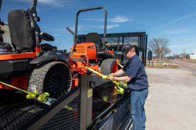 Flatbed truck loading Isuzu NQR gas engine with orange compact utility vehicle secured on trailer by worker tightening ratchet straps.