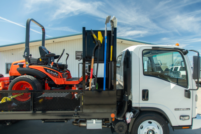 Isuzu NQR-GAS flatbed truck with a Kubota tractor on the flatbed carrying shovels and equipment visible in the truck bed.