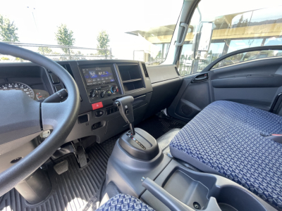 Isuzu NQR cab interior showing steering wheel, dashboard with digital radio display, climate controls, gear shifter, and blue patterned driver seat.