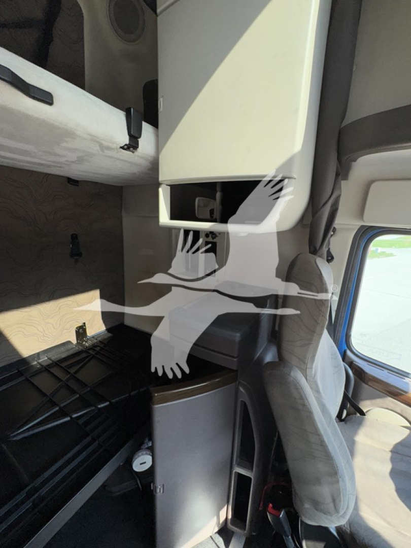 Cab interior of a Volvo VNL sleeper showing a storage compartment and lower cubby with a metallic door panel and stitching on the seat nearby.