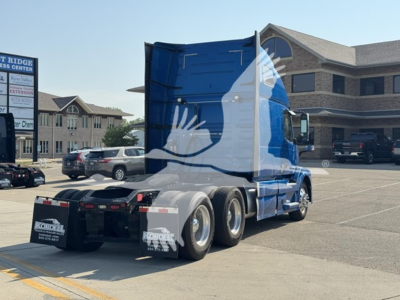 Semi truck rear and side view showing blue Volvo VNL 64T670 sleeper cab with multiple rear axles in a parking lot.