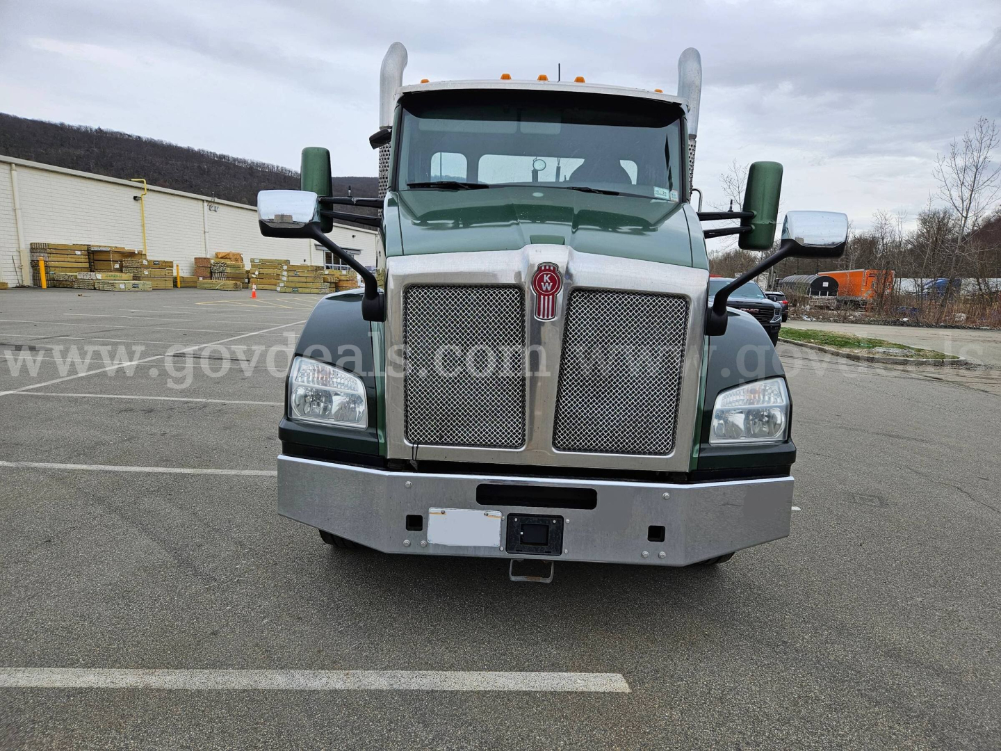 Kenworth T880 front view showing grille, bumper, and large rectangular headlights. Green cab with chrome mirrors and bumper, empty parking lot.
