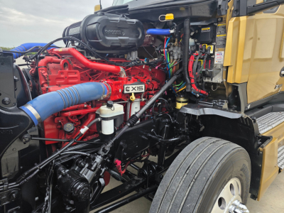 Kenworth T680 engine bay showing bright red Cummins X15 engine, blue intercooler piping, yellow cab, and black chassis with various hoses and wiring.