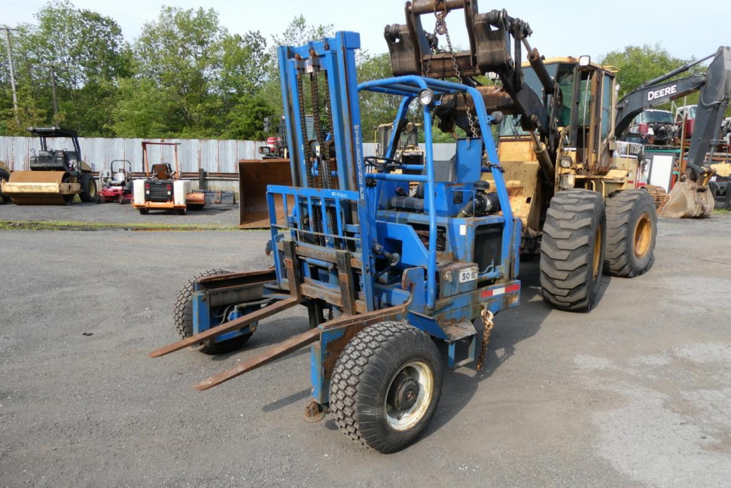 Teledyne D5000 forklift attachment in a yard with other heavy equipment; blue color, forks extended, vertical mast, single front wheel visible