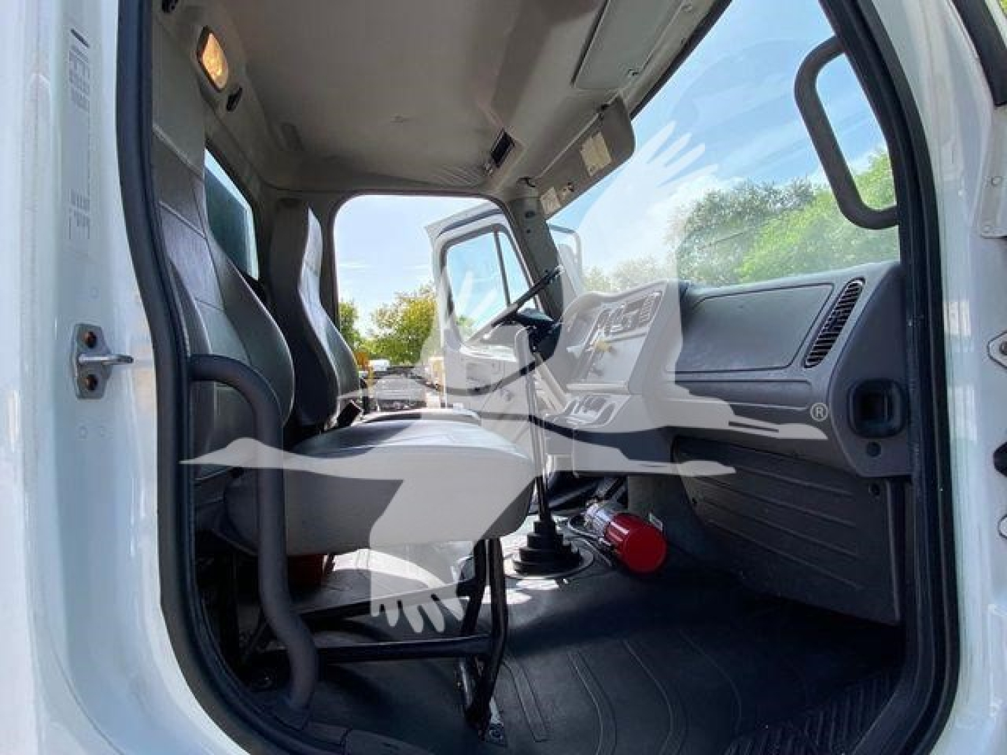 Truck cab interior view from driver side showing seating, steering wheel, dashboard, and gear shift lever. Cabin is uncluttered with a red handheld extinguisher mounted near floor.