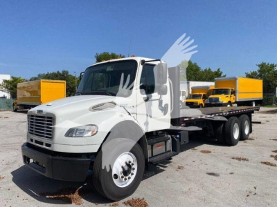 Freightliner M2 106 flatbed truck in white, front-left view, with yellow boxes in background.