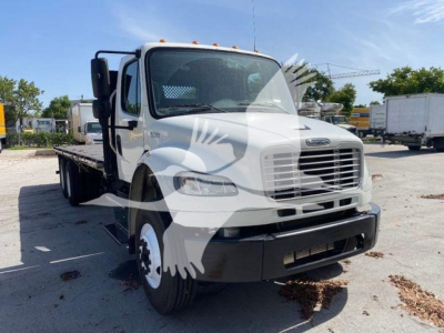 Flatbed truck in front view showing Freightliner M2-106 cab with white paint and black bumper. VIN label not visible. Flatbed chassis visible.