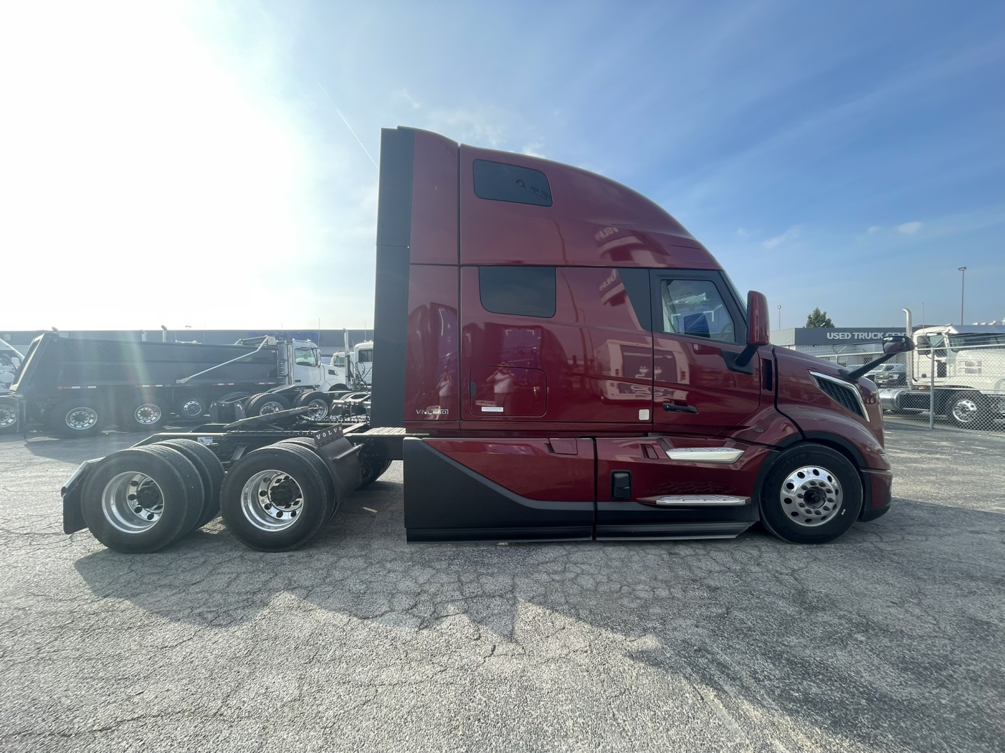 Side view of a maroon Volvo VNL860 conventional sleeper truck tractor with a full sleeper cab in a dealership lot. Visible text on the cab includes no legible slogans; appears to be new with triple rear axles and chrome exhaust, low front b